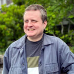 Dave Leo Baker - Musical Artist. Smiling middle aged man with short hair, wearing a grey jacket and striped jumper with tree in the background.